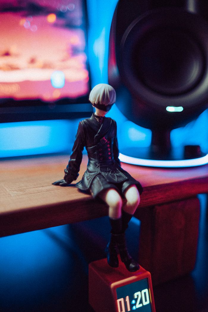 A stylish figurine sits on a desk lit by colorful neon lights with a digital clock display.